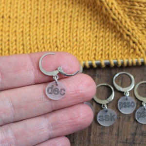 May include: Four clear plastic stitch markers with metal rings. The markers are labeled "dec", "ssk", "psso", and "psso".