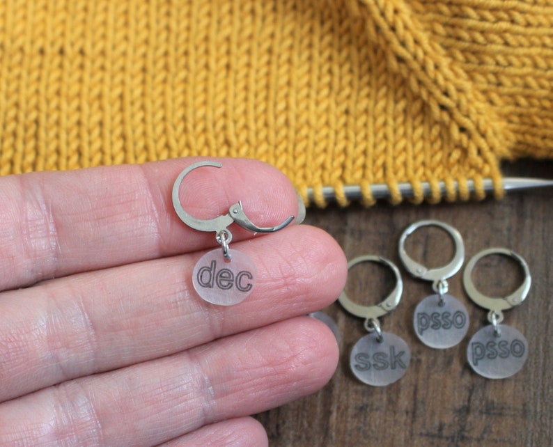 May include: Four clear plastic stitch markers with metal rings. The markers are labeled "dec", "ssk", "psso", and "psso".