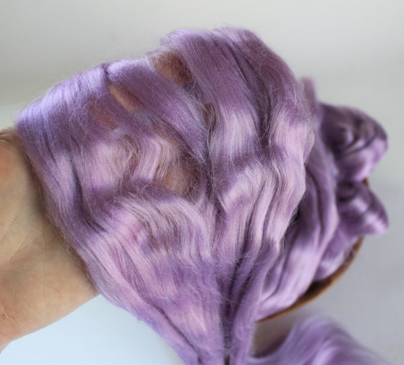Peut inclure: Gros plan sur une poign&eacute;e de fibres de couleur lavande. La fibre a une texture douce et ondul&eacute;e, ressemblant &agrave; de la soie ou &agrave; un mat&eacute;riau similaire. La couleur est un m&eacute;lange de violets clairs et fonc&eacute;s, cr&eacute;ant un effet de d&eacute;grad&eacute; subtil. La fibre est tenue dans une main.