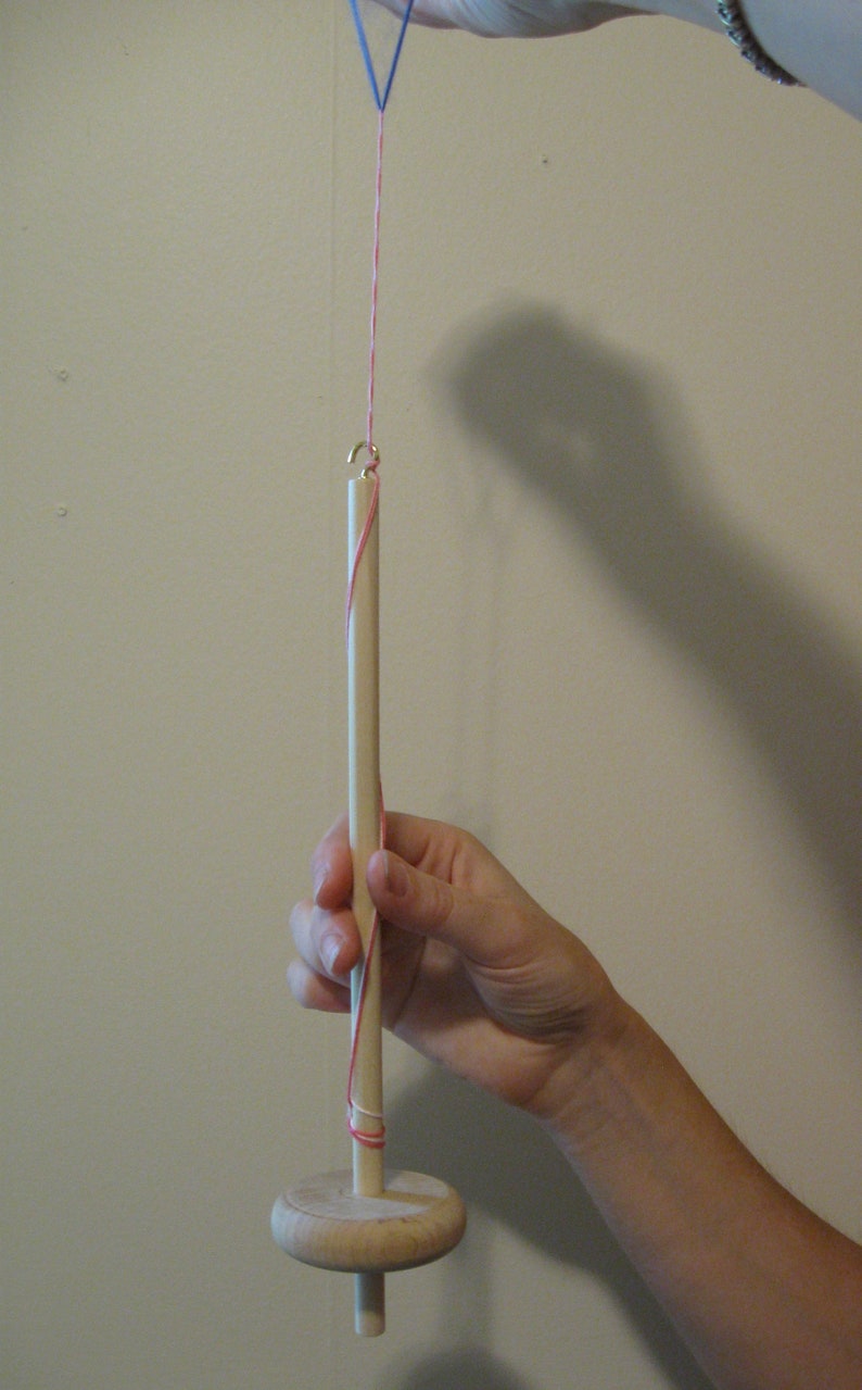 May include: A wooden spinning wheel with a red and blue string wrapped around it. The wheel is being held by a hand.