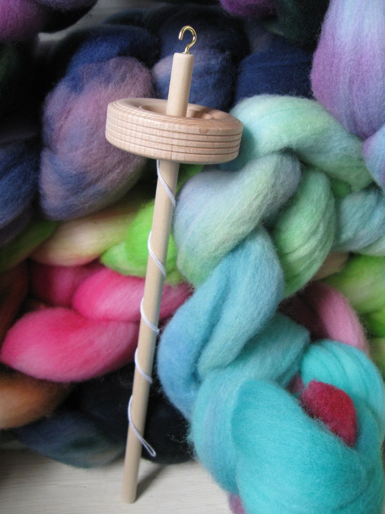 May include: A wooden spinning wheel with a gold hook and a white string wrapped around it. The spinning wheel is surrounded by colorful yarn.