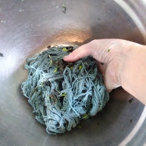 May include: A close-up of a bowl filled with a large clump of blue yarn. The yarn is tangled and has small green leaves mixed in.