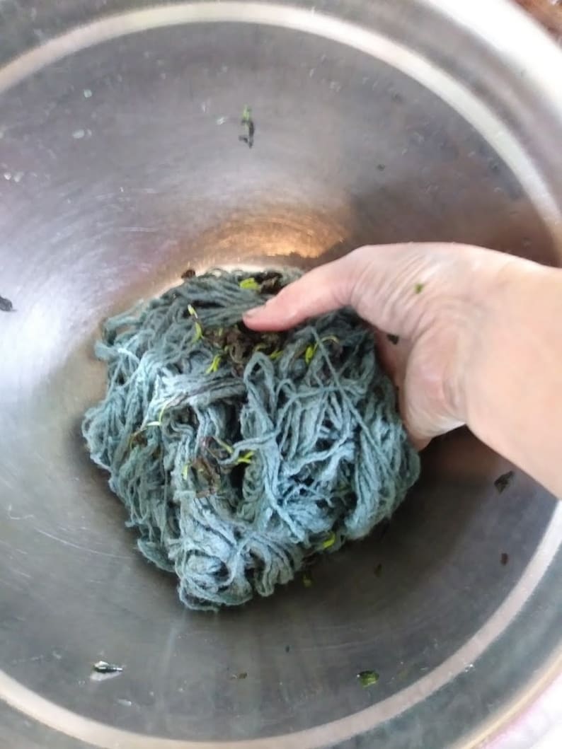May include: A close-up of a bowl filled with a large clump of blue yarn. The yarn is tangled and has small green leaves mixed in.
