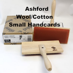 May include: A set of two Ashford wool and cotton hand carders. The carders are made of wood and have a red and a white surface. The carders are used for preparing wool and cotton for spinning.