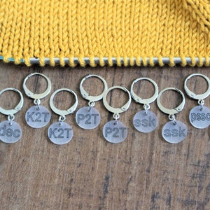 May include: Ten silver stitch markers with clear plastic beads. Each bead has a different knitting abbreviation printed on it: dec, K2T, P2T, ssk, psso.