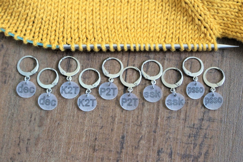 May include: Ten silver stitch markers with clear plastic beads. Each bead has a different knitting abbreviation printed on it: dec, K2T, P2T, ssk, psso.