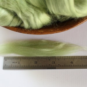 Puede incluir: Primer plano de una regla que mide un hilo de fibra de lana verde claro. La fibra mide aproximadamente 10 cm de largo.