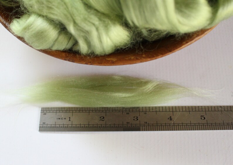 Puede incluir: Primer plano de una regla que mide un hilo de fibra de lana verde claro. La fibra mide aproximadamente 10 cm de largo.