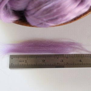 Peut inclure: Un &eacute;chantillon de fibre de couleur lavande, probablement de la laine ou de la soie, est pr&eacute;sent&eacute; &agrave; c&ocirc;t&eacute; d'une r&egrave;gle. La fibre mesure environ 10 cm de long, avec une texture douce et a&eacute;r&eacute;e. Une plus grande portion de la m&ecirc;me fibre est dans un bol en bois &agrave; l'arri&egrave;re-plan.