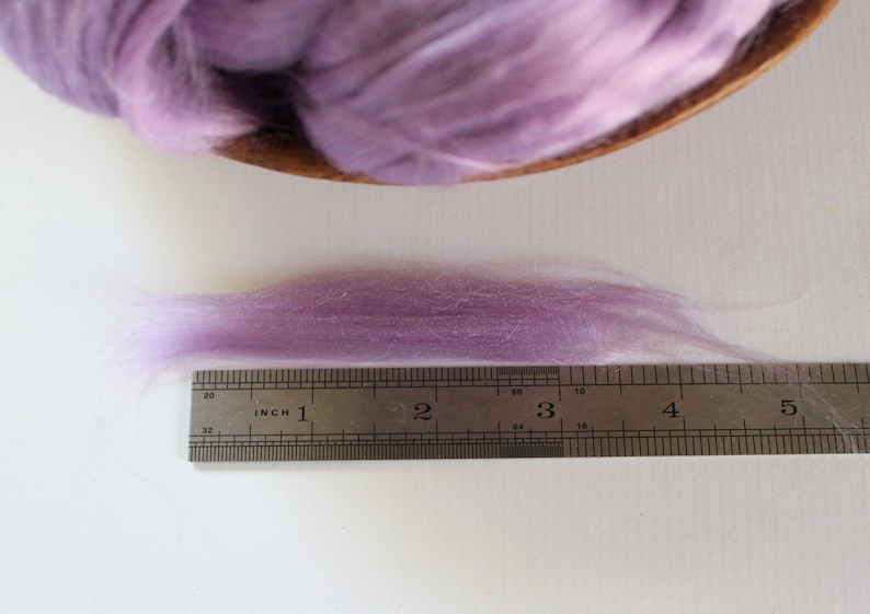 Peut inclure: Un &eacute;chantillon de fibre de couleur lavande, probablement de la laine ou de la soie, est pr&eacute;sent&eacute; &agrave; c&ocirc;t&eacute; d'une r&egrave;gle. La fibre mesure environ 10 cm de long, avec une texture douce et a&eacute;r&eacute;e. Une plus grande portion de la m&ecirc;me fibre est dans un bol en bois &agrave; l'arri&egrave;re-plan.
