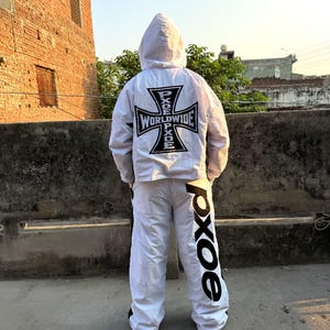 May include: A white hooded windbreaker jacket and matching white trousers set. The back of the jacket features a large black iron cross graphic with the text PXOE WORLDWIDE. The trousers have the text PXOE printed in large black letters.