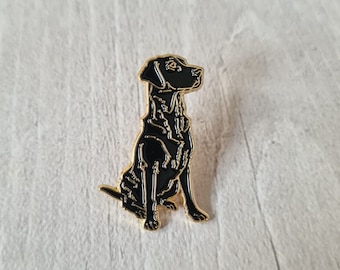 May include: A black Labrador Retriever enamel pin with gold-colored trim. The pin depicts a sitting dog with detailed features. The pin is set against a light wood grain background.