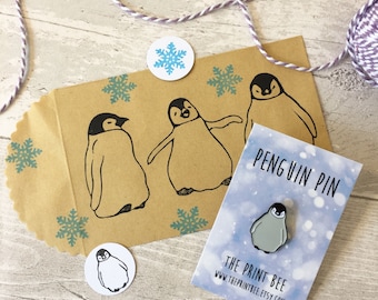 May include: A kraft paper gift bag with three black penguin illustrations and turquoise snowflakes. A small penguin pin on a card that reads "Penguin Pin" is also present, along with a round sticker with a penguin illustration.