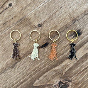 Puede incluir: Cuatro llaveros con forma de perro en varios colores: marr&oacute;n oscuro, blanco roto, marr&oacute;n claro y negro. Cada llavero tiene un anillo dorado. Los llaveros est&aacute;n colocados sobre una superficie de madera.