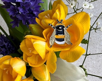 May include: A colorful enamel bee pin with black and yellow stripes, white accents, and silver-colored wings rests on a bed of yellow freesia flowers. The floral arrangement includes purple hyacinths, white tulips, and small white flowers.