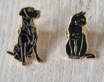 May include: Two enamel pins: a black Labrador Retriever and a black cat, both with gold-colored outlines. The dog pin is sitting, facing left. The cat pin is sitting, facing right. Both pins have a glossy finish.