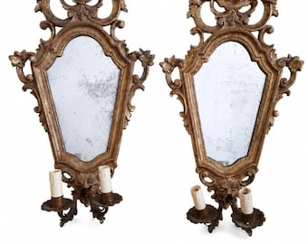 May include: A pair of gold-toned wall sconce mirrors featuring ornate, carved frames with scrolling details. Each mirror has a central glass panel and two candle-style light fixtures at the base with white cylindrical covers.