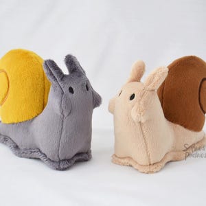 Peut inclure: Deux jouets en peluche en forme d'escargot. L'un est gris avec une coquille jaune, l'autre est beige avec une coquille marron. Les deux ont de petits yeux noirs et des oreilles droites. Les jouets sont doux et câlins.