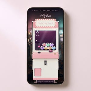 以下が含まれることがあります： ピンクと白のアーケードクレーンゲーム機のデジタルイラストが表示されたスマートフォン画面。機械には「A NIGHT AT THE ARCADE, FOR Sophie」「Catch my heart」「Drop the token in the slot」という文字と、内部にカラフルな球体が描かれています。