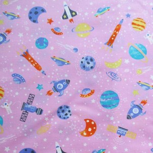 May include: Pink fabric with a repeating pattern of rockets, planets, stars, and spaceships in shades of blue, orange, yellow, and white. The design features various celestial bodies and spacecraft, creating a space-themed textile.