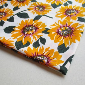 May include: White fabric with a repeating pattern of yellow sunflowers with pink and burgundy centers. The fabric is folded in the lower right corner.