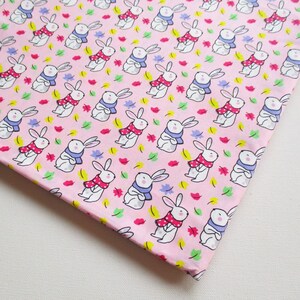 May include: Pink fabric with a repeating pattern of white bunnies wearing blue scarves and red polka dot scarves. The bunnies are surrounded by small green, yellow, and pink leaves.