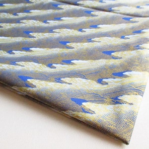 May include: Folded fabric displaying a repeating wave pattern in blue, gold, and grey. The design has a textured look, with overlapping waves creating movement. The fabric has a subtle sheen. The pattern is reminiscent of traditional Japanese art.