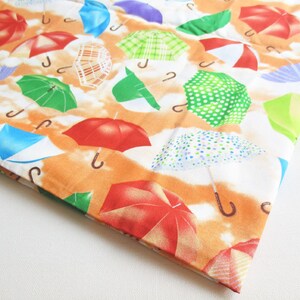 May include: A white fabric with a repeating pattern of colorful umbrellas. The umbrellas are in various colors, including red, blue, green, yellow, and purple. The background is a light orange color.