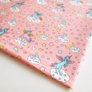 Puede incluir: Tela rosa con un patrón repetido de unicornios, nubes, estrellas, corazones y medias lunas. Los unicornios son de color azul claro y rosa con detalles de arcoíris.