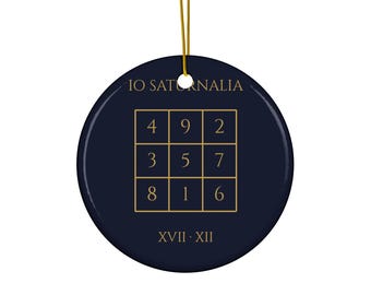 Könnte beinhalten: Ein rundes, dunkelblaues Keramikornament mit einem goldenen magischen Quadratgitter mit den Zahlen 1 bis 9. Der Text IO SATURNALIA steht über dem Gitter und XVII · XII darunter. Es hängt an einer dünnen, goldfarbenen Kordel.