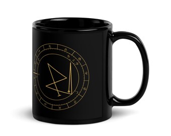 Könnte beinhalten: Ein schwarzer Keramikbecher mit einem goldfarbenen, kreisförmigen astrologischen Diagramm. Die Grafik zeigt ein zentrales geometrisches Siegel, das von einem Ring aus Tierkreiszeichen und Planetenmarkierungen in feinen Goldlinien umgeben ist.