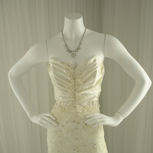 Può includere: Un abito da sposa bianco senza spalline in raso e pizzo con un corpetto decorato con perline e una gonna aderente.