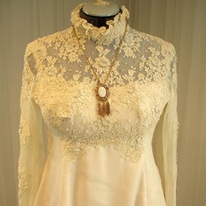 Könnte beinhalten: Ein weißes Spitzen-Hochzeitskleid mit langen Ärmeln und einem hohen Ausschnitt. Das Kleid hat ein zartes Blumenmuster und ist mit einer goldenen Anhänger-Halskette verziert.