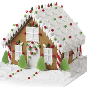 May include: A gingerbread house decorated with white frosting, red and green candies, and candy canes. The house has a white door with a red and green candy wreath. There are two green gumdrop trees in front of the house.