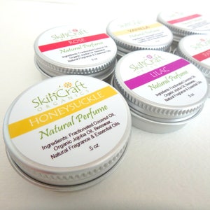 May include: Six small silver tins with white labels and colorful accents. The labels read "SkinCraft Organics" and list different scents: Rose, Vanilla, Lilac, Patchouli, Honeysuckle. Each tin contains 5 oz of natural perfume.