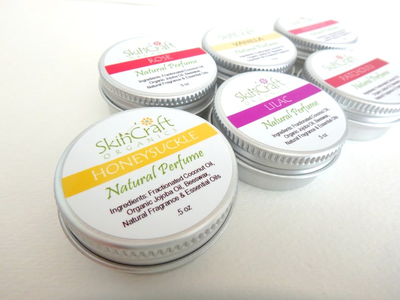 May include: Six small silver tins with white labels and colorful accents. The labels read "SkinCraft Organics" and list different scents: Rose, Vanilla, Lilac, Patchouli, Honeysuckle. Each tin contains 5 oz of natural perfume.