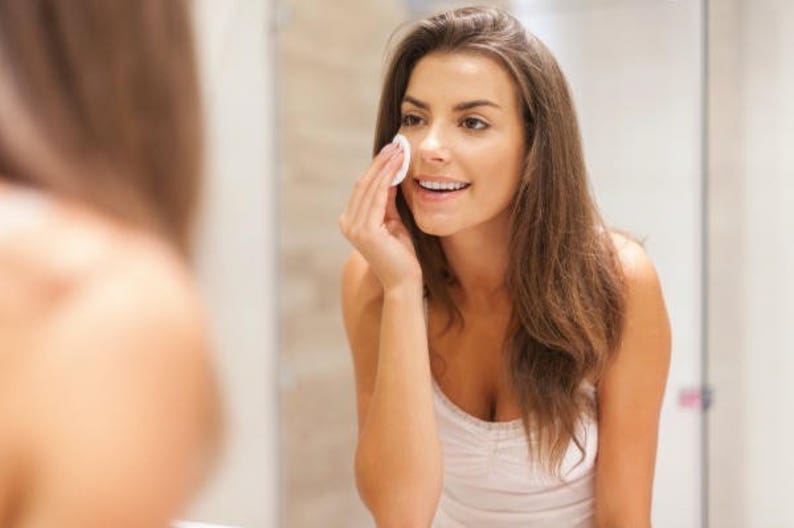 May include: A woman with long brown hair is applying a cotton pad to her face in front of a mirror. She is wearing a white vest top.