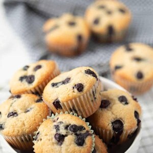 Pu&ograve; includere: Una ciotola bianca piena di muffin ai mirtilli appena sfornati. I muffin hanno la parte superiore dorata e sono cosparsi di mirtilli scuri. Alcuni muffin sono sullo sfondo ed &egrave; visibile un panno grigio.