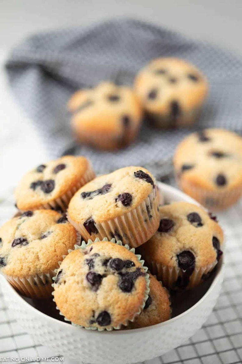 Pu&ograve; includere: Una ciotola bianca piena di muffin ai mirtilli appena sfornati. I muffin hanno la parte superiore dorata e sono cosparsi di mirtilli scuri. Alcuni muffin sono sullo sfondo ed &egrave; visibile un panno grigio.