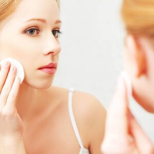 May include: A woman with long, red hair is applying a cotton pad to her face in front of a mirror.