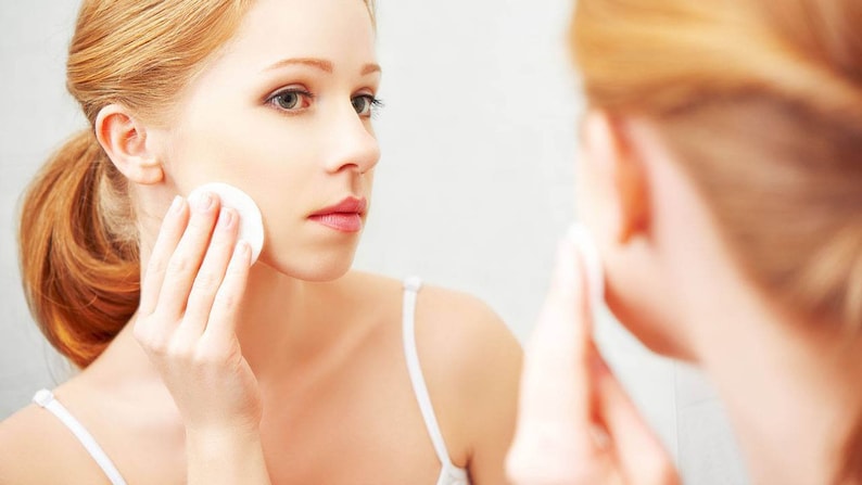 May include: A woman with long, red hair is applying a cotton pad to her face in front of a mirror.
