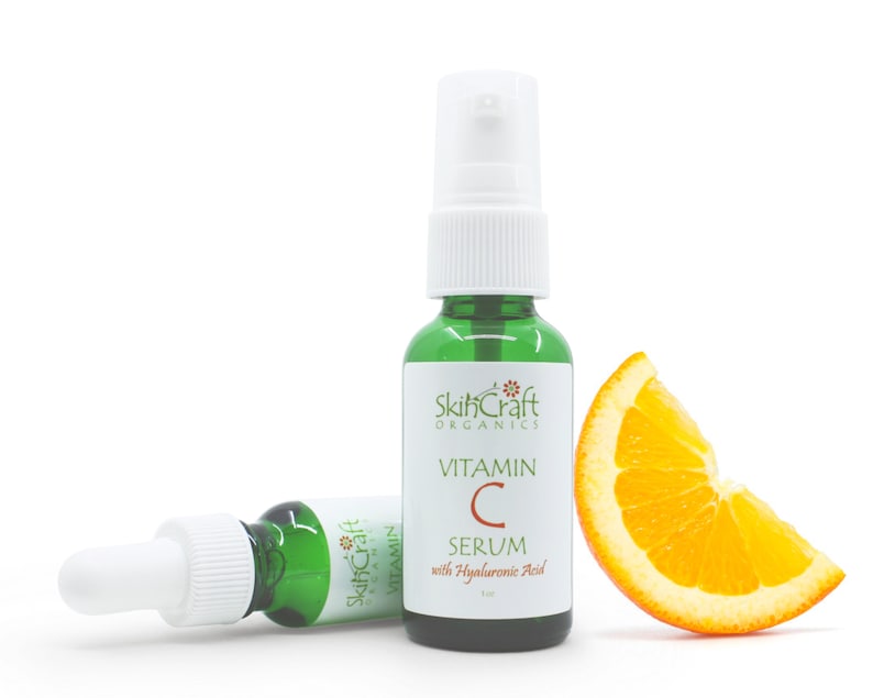 May include: Two green glass bottles of SkinCraft Organics Vitamin C Serum with Hyaluronic Acid. The bottle on the right has a white pump top. The bottle on the left has a white dropper top. A slice of orange is in the foreground.