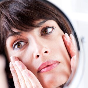 May include: A woman looks in a mirror and touches her face with both hands. She has brown hair and is wearing a white shirt.