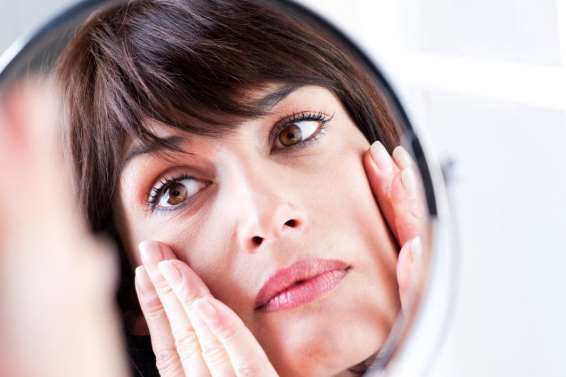 May include: A woman looks in a mirror and touches her face with both hands. She has brown hair and is wearing a white shirt.