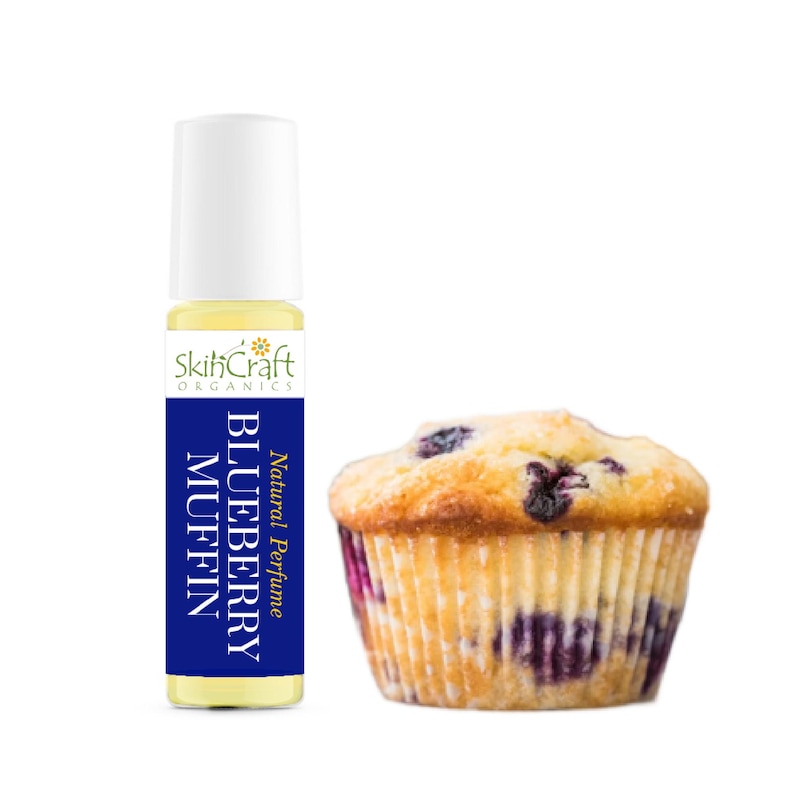 Pu&ograve; includere: Un flacone di profumo roll-on con tappo bianco ed etichetta blu con la scritta "BLUEBERRY MUFFIN" e "Profumo Naturale". Accanto, un muffin ai mirtilli cotto in un pirottino di carta. Il flacone di profumo ha il marchio "SkihCraft Organics".