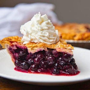 Pu&ograve; includere: Una fetta di torta ai mirtilli su un piatto bianco, guarnita con panna montata. La torta ha una crosta dorata e un ripieno di mirtilli blu scuro. Un'altra torta &egrave; visibile sullo sfondo.