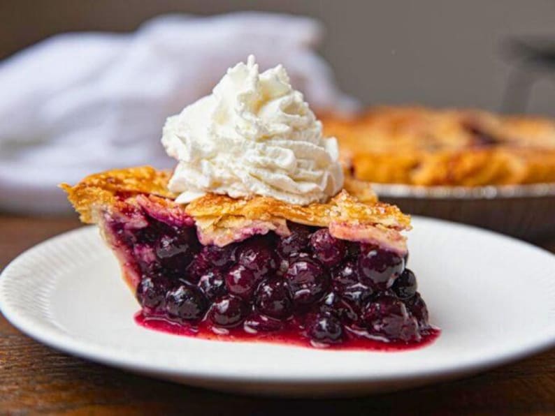 Pu&ograve; includere: Una fetta di torta ai mirtilli su un piatto bianco, guarnita con panna montata. La torta ha una crosta dorata e un ripieno di mirtilli blu scuro. Un'altra torta &egrave; visibile sullo sfondo.