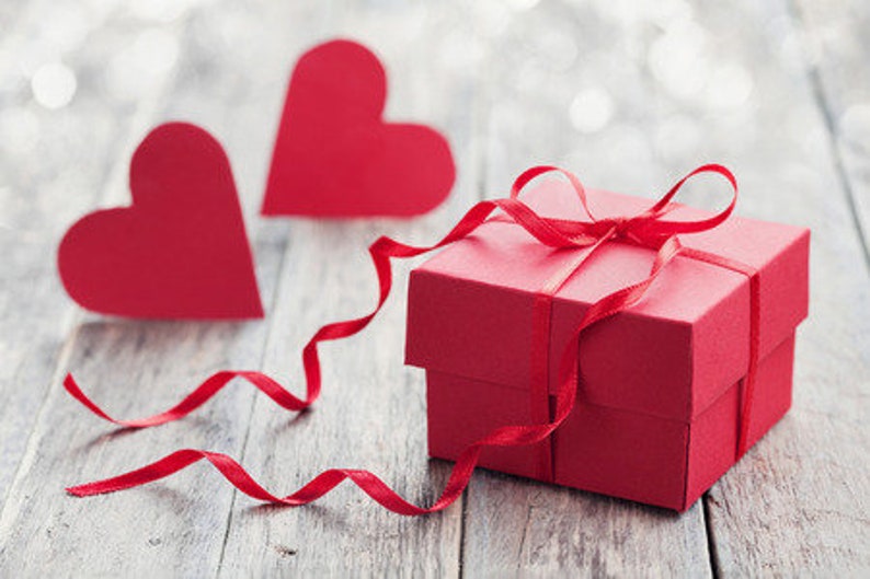 May include: A red gift box with a red ribbon tied in a bow. Two red paper hearts are on a wooden surface in the background.