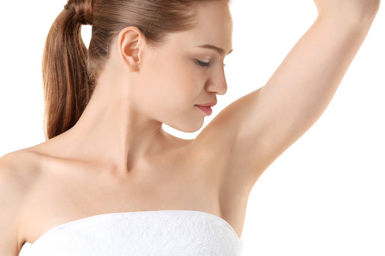 May include: A woman with long brown hair pulled back in a ponytail, wearing a white strapless top, raises her left arm to show her underarm.