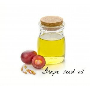 May include: A glass bottle with a cork stopper filled with yellow grape seed oil. Two halved red grapes and grape seeds are next to the bottle. The text "Grape seed oil" is written in cursive below the bottle.